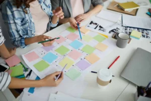 Personas colaborando en la planificación de un proyecto con notas adhesivas de colores en un tablero, rodeadas de papeles, lápices y una taza de café sobre una mesa de trabajo.