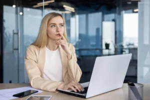 Mujer joven pensando frente a una computadora portátil en una oficina moderna.