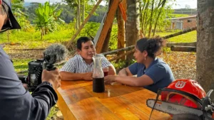 Grabación de video en finca rural: pareja conversa en mesa de madera con cámara y equipo Husqvarna al frente.